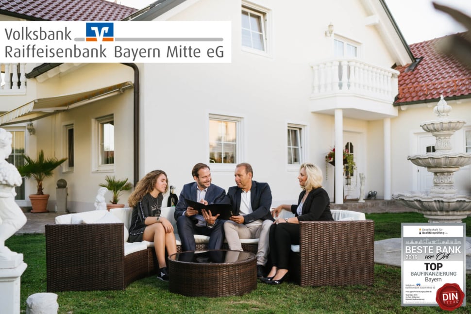 Ein Baufinanzierungsberater sitzt im Garten vor einem Immobilie mir einer glücklichen Familie